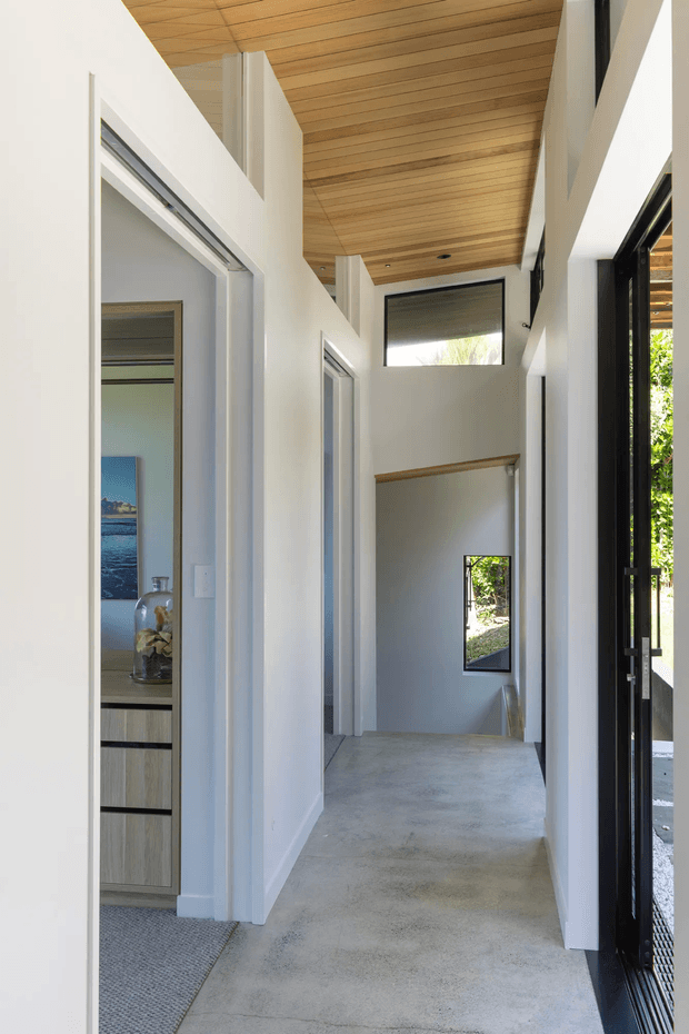 A bright contemporary residential hallway and stair landing with white walls, polished concrete flooring, a warm timber-lined ceiling, and black-framed glazing and entry doors. The corridor opens toward a stairwell with a small window and timber handrail, while a partially visible room with built-in light timber cabinetry and artwork appears to the left. The space has a tall, airy volume and strong natural light.