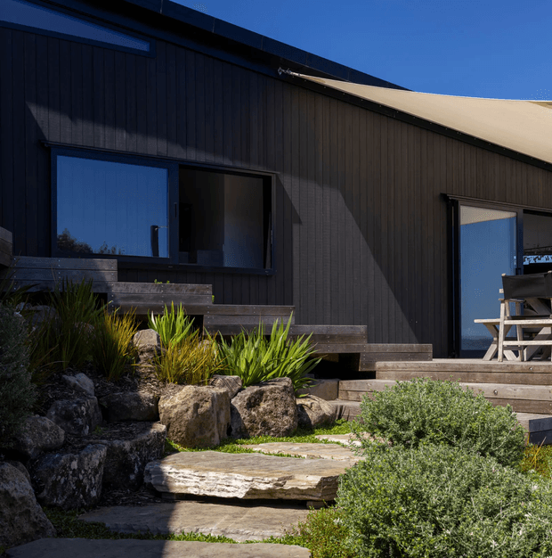 Exterior view of a dark-clad, single-storey dwelling with a sloping roof, large glazed openings, and timber steps leading to a raised deck. A naturalistic rock garden and layered stone pathway occupy the foreground, while outdoor seating is visible on the deck.