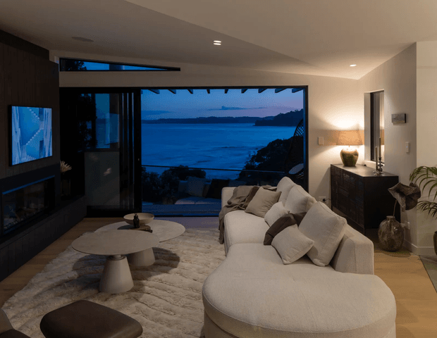 A refined contemporary living room with a large sectional sofa arranged around low sculptural coffee tables and a wall-mounted television. Full-height glazing opens to a blue waterfront or coastal view, while dark built-in media cabinetry contrasts with warm neutral walls, timber-toned flooring, textured rug, and soft layered lighting. A dark sideboard and indoor plants add warmth to the elegant, relaxed space.