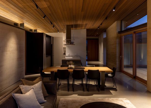 Warm, moody open-plan dining and kitchen interior with a long timber dining table, dark dining chairs, compact kitchen cabinetry and island, exposed timber ceiling, concrete-toned walls, and extensive timber-framed glazing. A living area is partially visible in the foreground, while a hallway extends beyond the kitchen.