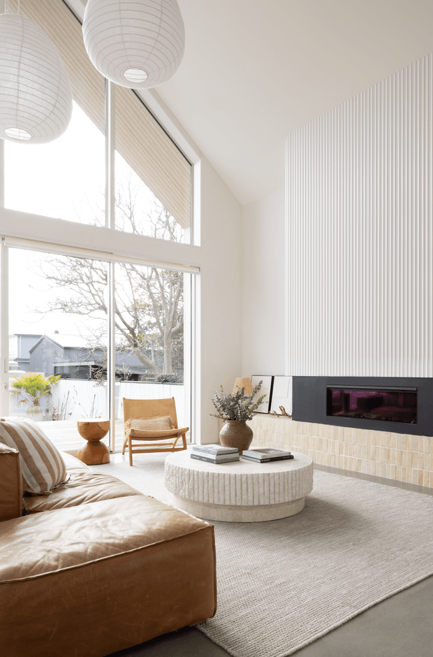 Bright contemporary living room with a double-height feel, expansive floor-to-ceiling glazing, white ribbed wall panelling, and a long black fireplace set above a pale textured tile surround. The room features a tan leather sectional, sculptural round coffee table, woven rug, timber lounge chair, ceramic vessel, and large white pendant lights. A suburban exterior is visible through the windows.
