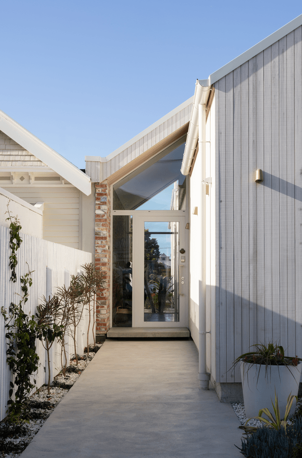 A narrow contemporary residential entry passage leading to a glazed front door. The facade combines pale vertical cladding, white weatherboard siding, exposed red brick, a sloping roofline, white gutters, and a concrete walkway bordered by white fencing and planted beds.