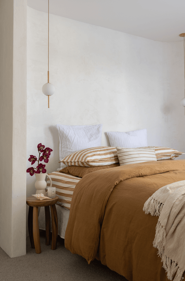 A warm, softly styled bedroom with a low-profile bed dressed in ochre-brown linen bedding, striped beige-and-white pillows, and a fringed cream throw. The room has textured off-white plaster walls, neutral carpet, sculptural globe pendant lights with timber or brass-toned details, and a small dark timber side table topped with a vase of burgundy flowers.