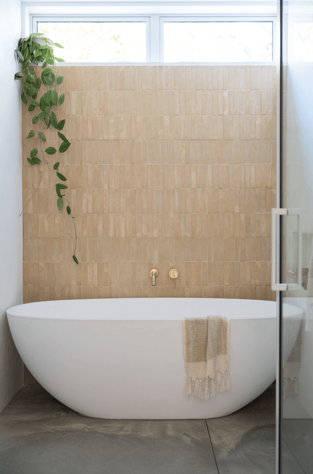 Bright contemporary bathroom featuring a freestanding white oval bathtub, a full-height beige vertically textured tile feature wall, wall-mounted bath fittings, large frosted or bright windows, a trailing indoor plant, and a glass shower screen at the right. The floor has a grey concrete-like finish and the overall palette is calm and natural.