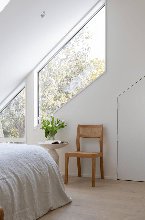 A serene, minimalist bedroom corner with a low-profile bed dressed in white linen, pale timber flooring, and a simple woven timber chair beside a sculptural side table. Large angular windows bring in abundant natural light and frame leafy trees outside. The room has crisp white walls, a clean flush door, and understated contemporary detailing.