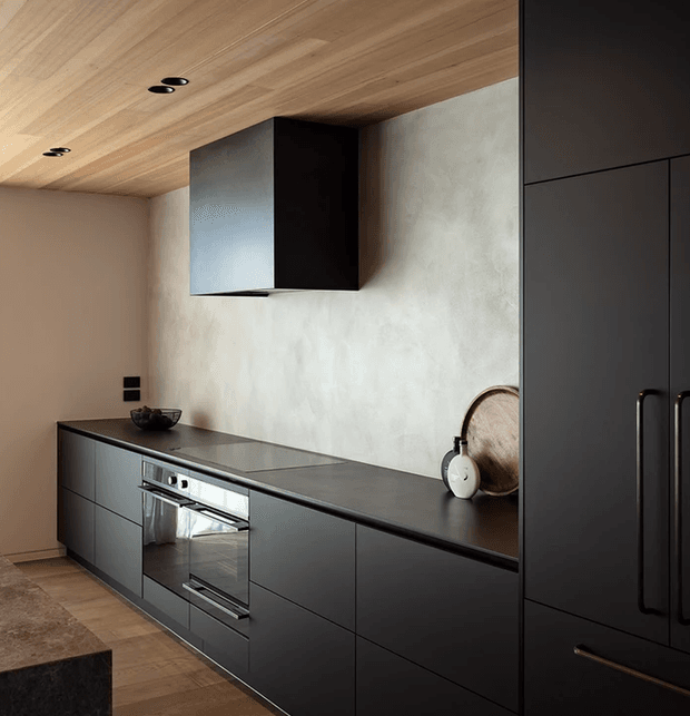 A refined contemporary kitchen with full-height matte dark cabinetry, an integrated oven and cooktop, a slim dark benchtop, and a pale textured feature wall. The ceiling is lined with warm timber boards and recessed downlights, creating a warm contrast against the charcoal joinery. Minimal decorative accessories and clean horizontal lines give the space a sleek, understated appearance.