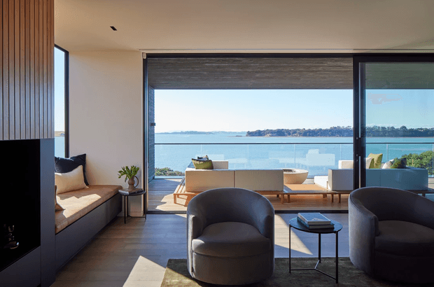A refined modern living room with floor-to-ceiling glazing overlooking a calm waterfront. The room features dark timber flooring, a built-in window seat, sculptural upholstered lounge chairs, a low coffee table, a linear fireplace wall with vertical timber battens, and a neutral sofa arrangement facing the view. The palette is warm and restrained, with strong indoor-outdoor connection and abundant natural light.