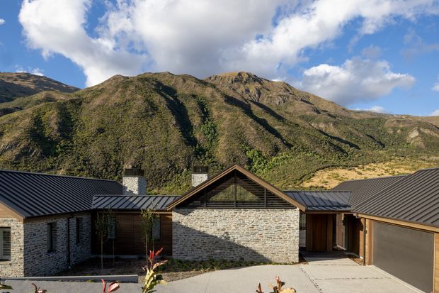 Exterior view of a low-rise mountain home arranged around a paved central courtyard. The building has dark standing-seam pitched roofs, pale stacked-stone walls, timber-framed glazed sections, and a central gabled volume with a high triangular window. Rugged green-brown mountains rise immediately behind the property under a partly cloudy blue sky.
