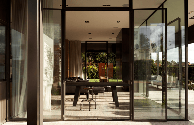 A refined contemporary indoor-outdoor living and dining area viewed through expansive black-framed glass doors. The space features a long dark dining table with stools, a sofa and lounge seating beyond, warm timber or wood-toned curtains and surfaces, polished light flooring, and strong visual connection to a planted outdoor terrace. Abundant glazing and sunlight create a calm, airy atmosphere with crisp linear detailing.
