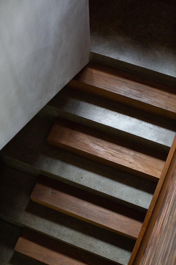 Close-up view looking down a narrow interior stairway with floating timber treads set into a dark, rough concrete stair structure. A plain light plaster wall borders the left side, while a timber handrail or balustrade runs along the right. The composition is subdued and materially focused, highlighting the contrast between warm wood and grey concrete.