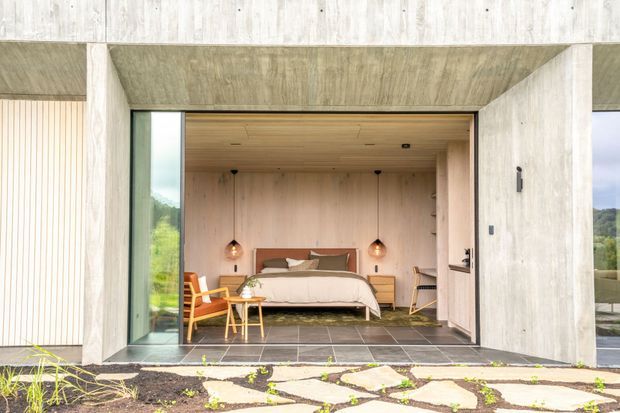 A minimalist modern bedroom viewed through a large open glazed portal in a concrete-framed facade. The room features light timber wall and ceiling lining, a bed with a warm wood headboard, compact bedside tables, pendant lights, a small desk and chair, and dark stone or tile flooring. The exterior includes pale raw concrete, vertical timber cladding, expansive glazing, and an irregular stone-paved garden path.
