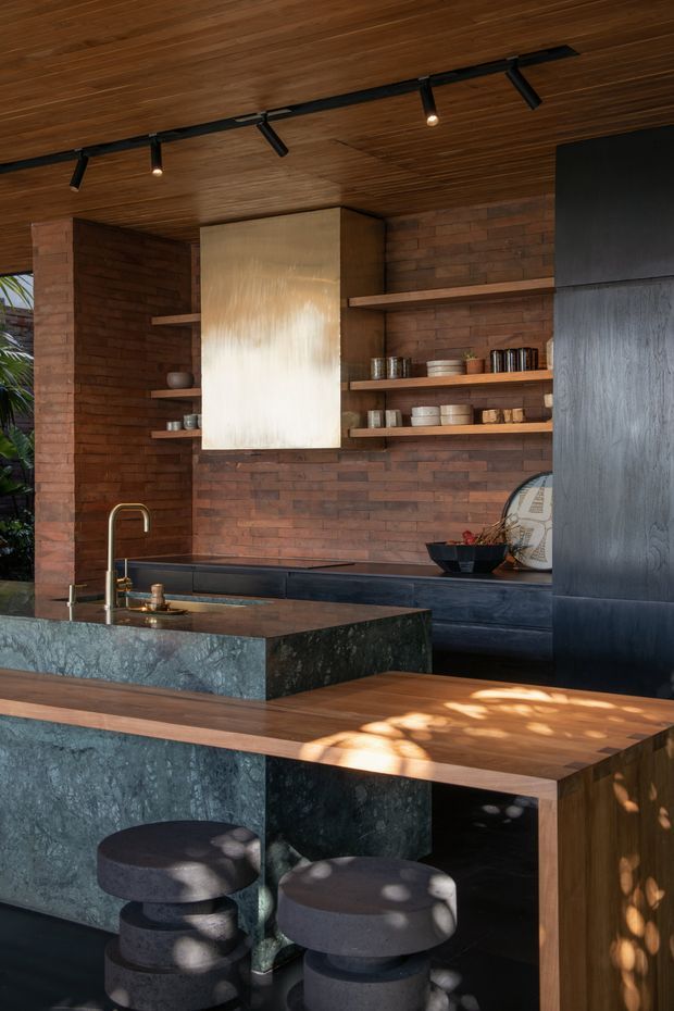 A sophisticated open-plan kitchen with dark green stone islands, warm timber cabinetry and ceiling panelling, exposed red-brown brick walls, and matte black detailing. Open timber shelves display ceramics and glassware, while a sculptural brushed-metal rangehood forms a central feature. Track lighting and dappled sunlight create a warm, contemporary atmosphere, with rounded dark upholstered stools at the island.
