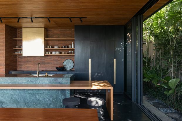 A sophisticated modern kitchen with a large stone island, dark timber and brick finishes, matte black full-height cabinetry, brass-toned handles, open shelving, and floor-to-ceiling glazing opening toward dense greenery. A timber-lined ceiling and track lighting add warmth and texture.