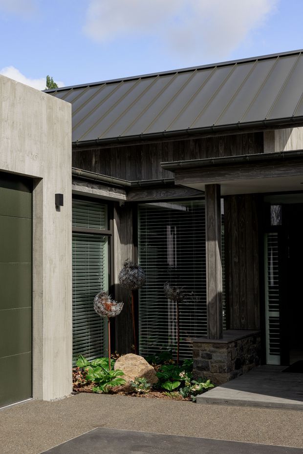 Exterior view of a contemporary residential entry courtyard. The facade combines dark vertical timber cladding, pale textured concrete walls, a standing-seam metal pitched roof, and large windows with horizontal blinds. A covered entry porch, stone-clad low wall, paved ground, and a small planted garden with rocks define the approach.