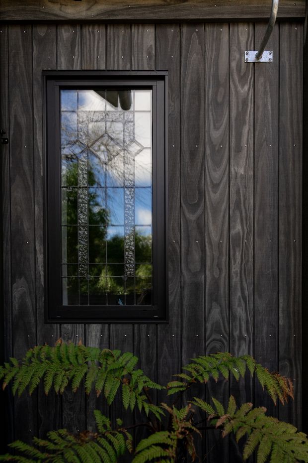 Close-up exterior detail of a dark, vertically boarded timber-clad wall with a black-framed decorative leadlight-style window. The window reflects blue sky and surrounding greenery, while lush ferns grow along the base. A simple metal wall-mounted fixture is visible at upper right. The image emphasises textured weathered timber, dark framing, and natural planting.