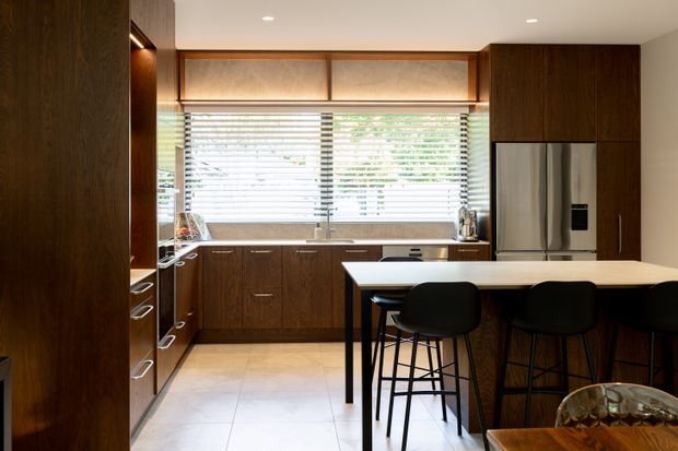A warm contemporary kitchen with dark timber cabinetry, a large island with black bar stools, stainless-steel refrigerator, pale tiled flooring, and wide windows fitted with horizontal blinds. Integrated warm lighting highlights the timber joinery, while a partial dining area is visible in the foreground.
