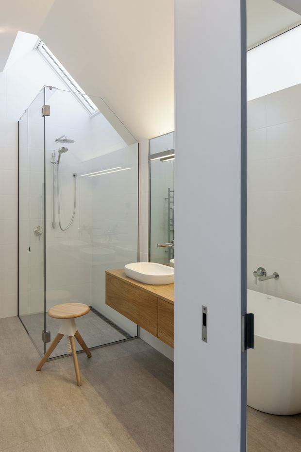 Bright contemporary bathroom with a large frameless glass walk-in shower, wall-mounted timber vanity with a white vessel basin, mirrored cabinetry, and a freestanding white bathtub partially visible behind an open door. A high angled skylight or clerestory brings in abundant natural light. The floor is finished in large warm grey tiles, with white tiled walls and pale neutral surfaces.