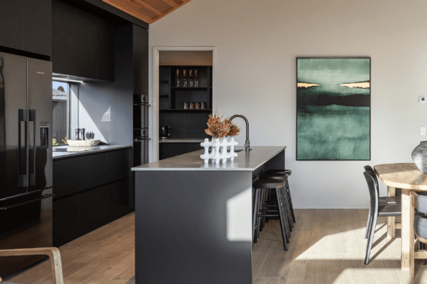 Contemporary open-plan kitchen and dining area with dark matte cabinetry, a central island with integrated sink and black stools, timber flooring, and a light neutral wall. A built-in display niche, timber ceiling detail, artwork, and a partially visible dining table complete the warm, refined interior. Strong sunlight creates contrasting geometric shadows across the island and floor.