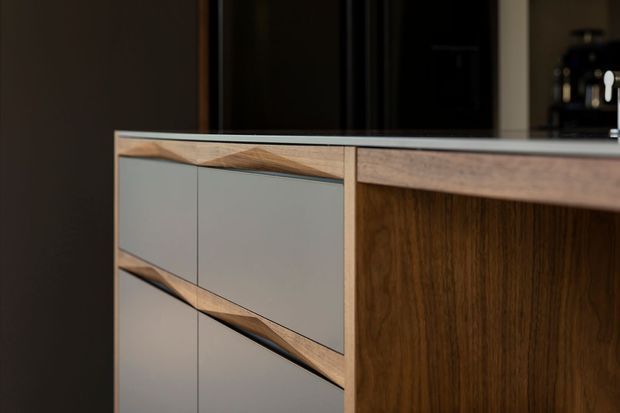 Close-up detail of a contemporary built-in kitchen or joinery unit, featuring warm walnut-toned timber panels, matte light-grey drawer fronts, and distinctive angled timber edging and pulls. A dark interior kitchen setting is visible in the background.