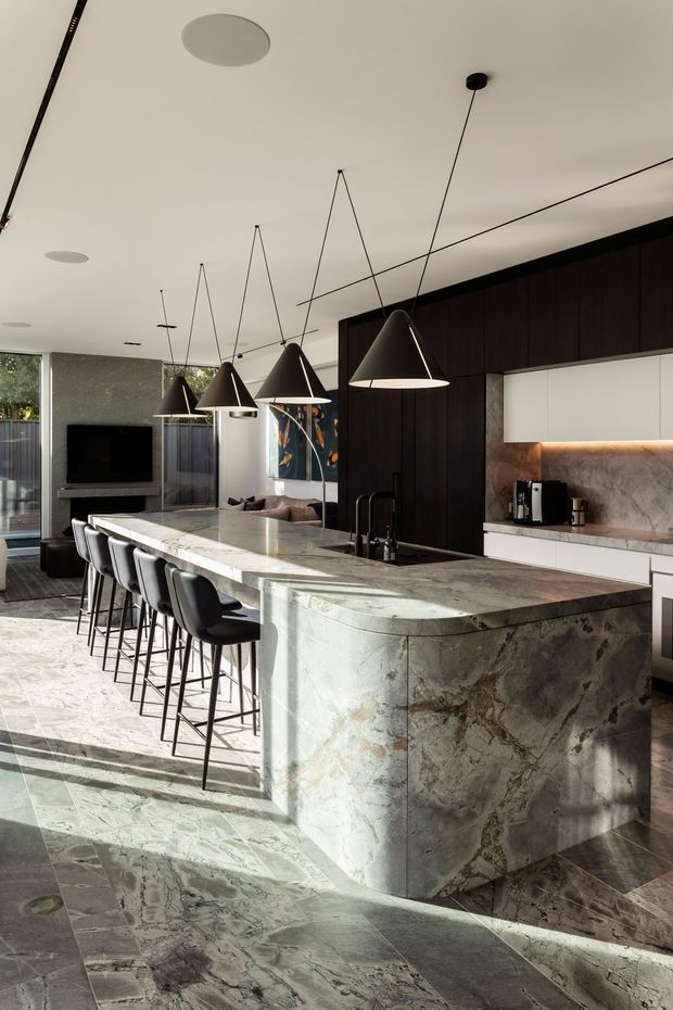 A sophisticated contemporary kitchen dominated by a large waterfall island clad in dramatic veined natural stone, with matching stone flooring and benchtop surfaces. Dark timber cabinetry forms a full-height wall, paired with a pale stone backsplash and warm under-cabinet lighting. Several black cone-shaped pendant lights hang above the island, which includes seating for multiple people and an integrated sink. A living area with a television is visible beyond the kitchen.