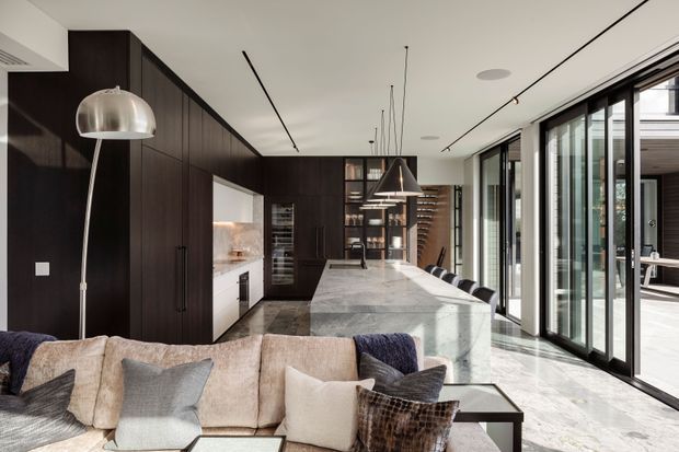 A sophisticated open-plan kitchen, dining and living space with dark timber cabinetry, a large pale stone island, full-height black-framed glazing, and a neutral sectional sofa in the foreground. Linear track lighting, pendant lights and contemporary built-in joinery create a refined, high-contrast interior with strong indoor-outdoor connection.