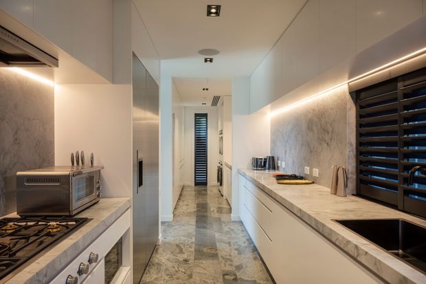A narrow, high-end galley kitchen with extensive white cabinetry, marble-look benchtops and splashback, integrated appliances, a gas cooktop and oven on the left, and a large sink beneath dark horizontal window louvres on the right. Warm linear under-cabinet lighting, polished stone flooring, and reflective appliance or cabinetry surfaces create a sleek contemporary fit-out. A hallway is visible beyond the kitchen.