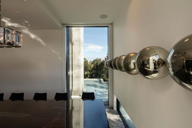 A minimalist dining area viewed along a dark glossy dining table toward a full-height glazed opening. Black dining chairs line the table, while a sculptural row of reflective spherical pendant lights dominates the right side. The pale walls and ceiling, narrow dark window framing, polished surfaces, and strong natural light create a clean, contemporary interior with an outdoor terrace and greenery visible beyond the glazing.