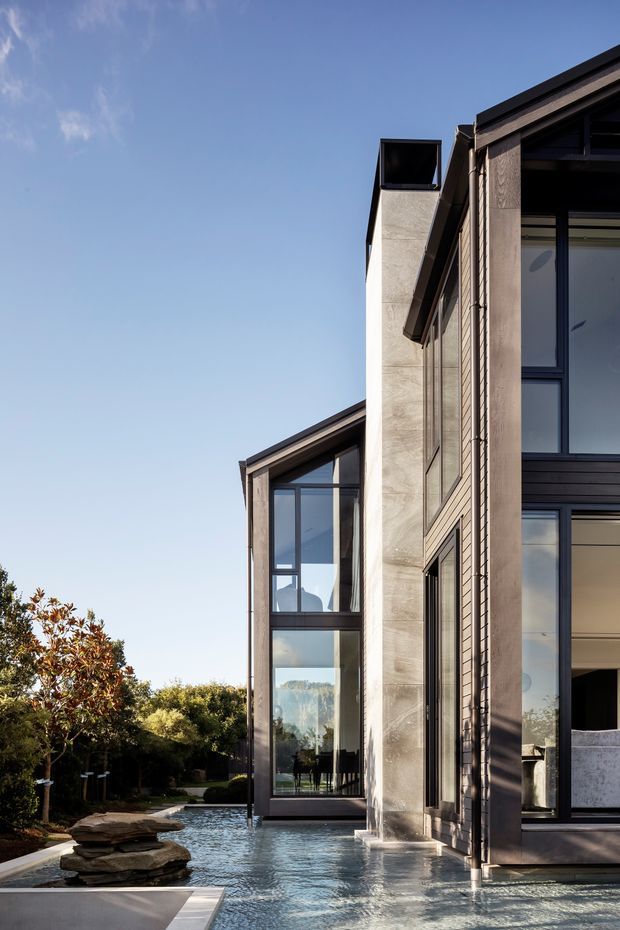 Exterior architectural view of a contemporary multi-level residence with tall dark-framed glazing, textured light-grey masonry or concrete walls, and dark roof edges. A shallow reflecting pool borders the house, with layered stone landscaping and mature planting visible at ground level. The image is photographed in clear daylight from outside, emphasizing the vertical facade composition and connection between the building and water feature.