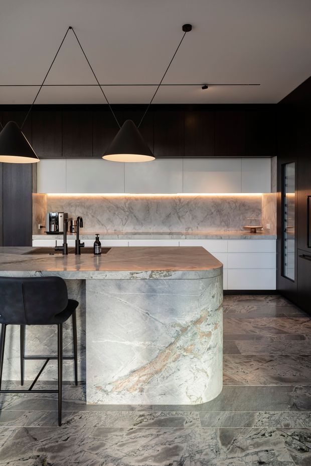 A sophisticated contemporary kitchen featuring a large waterfall island clad in heavily veined natural stone, dark timber cabinetry, pale white upper cabinetry, and a matching stone backsplash with warm integrated LED lighting. Matte black pendant lights, tapware, appliances, and bar stools provide contrast. The kitchen has a polished, high-end appearance with a generous island and dark patterned stone flooring.
