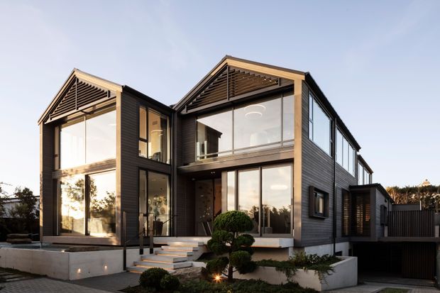 A contemporary multi-level residential house photographed from the front at dusk, featuring dark horizontal cladding, extensive floor-to-ceiling glazing, projecting gabled roof forms with exposed timber louvre detailing, and concrete entry terraces and planters. Carefully clipped shrubs and warm interior lighting create a refined modern street-facing facade.