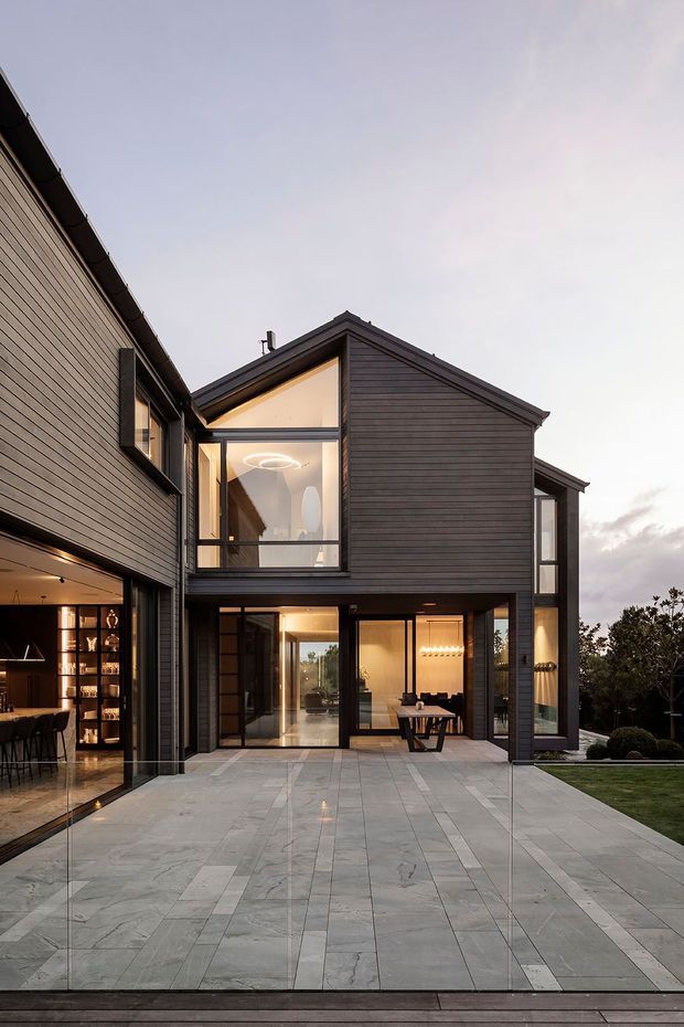 Exterior view of a contemporary multi-storey home at dusk, with dark horizontal cladding, steep gabled roof forms, extensive floor-to-ceiling glazing, and a large pale stone-paved terrace. Warm interior lighting reveals a kitchen/bar area, dining space, and living areas through the glazed openings. Landscaped lawn and planting sit beside the terrace.