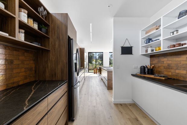 A contemporary galley kitchen with dark timber cabinetry, open shelving, a black stone benchtop, and warm brown brick-look splashback tiles. Light timber flooring and white walls create a bright, elongated space, with stainless-steel appliances and a bright windowed area at the far end.