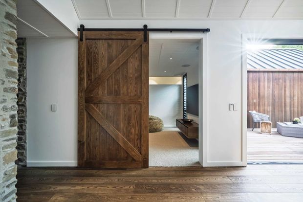 Bright contemporary interior focused on a large rustic timber barn-style sliding door in a white hallway or living area. The room has dark timber flooring, white painted walls, and a paneled white ceiling. A rough stacked-stone wall is visible at the left, while an opening reveals a carpeted sitting area and a glazed door leading to a timber deck and outdoor courtyard. The composition highlights the contrast between traditional timber and stone elements and clean contemporary finishes.
