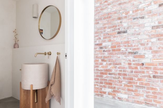 A compact, softly styled bathroom viewed through an open doorway, featuring a freestanding cylindrical vessel basin on a timber pedestal, wall-mounted brushed brass tapware, a round brass-framed mirror, pale textured wall tiling, and a white wall sconce. A red-toned exposed brick wall is visible immediately outside or adjacent to the bathroom.