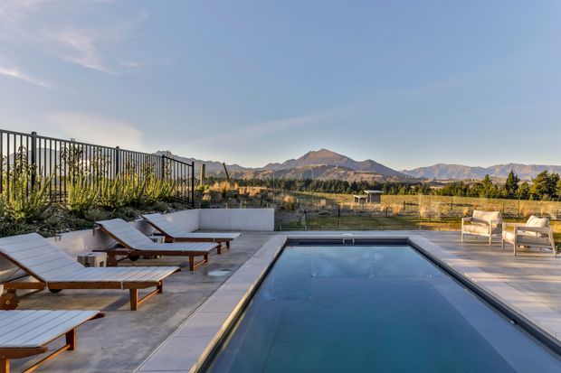 A modern outdoor pool terrace with a long rectangular swimming pool, timber-framed loungers, pale paving, metal railings, and a small seating area. The property overlooks a broad rural valley and rugged mountain range beneath a clear blue sky.