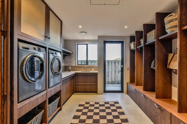 A well-appointed laundry room with a warm, dark timber fit-out. Full-height cabinetry houses stacked front-loading washing and drying appliances on the left, while open shelving and drawers provide storage on the right. A compact timber-toned benchtop and sink sit beneath a window, with a glazed exterior door, recessed ceiling lights, and a checkerboard rug completing the space.
