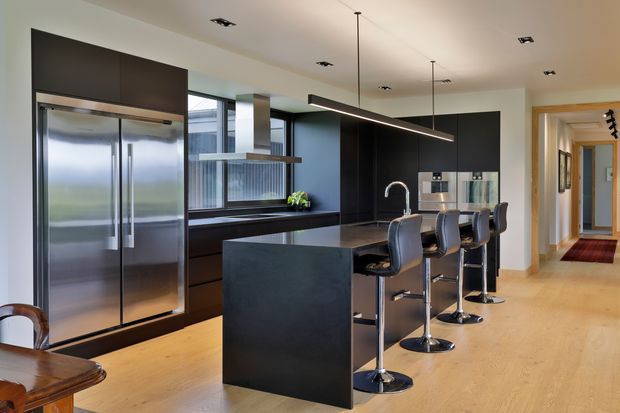 A spacious contemporary kitchen with dark flat-panel cabinetry, stainless-steel appliances, a large central island with a dark stone-like benchtop, and four bar stools. Warm timber flooring contrasts with the black joinery and pale walls. Integrated strip pendant lighting, recessed ceiling spotlights, and a hallway extending beyond the kitchen are visible.