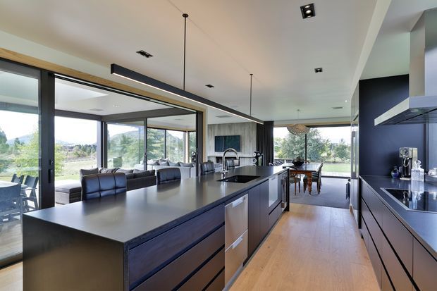 A spacious contemporary open-plan kitchen with a large dark island, integrated appliances, dark fixed cabinetry, and a contrasting timber floor. Floor-to-ceiling glazing connects the kitchen to an outdoor setting, while the adjacent dining and living areas are visible beyond. Recessed ceiling lights and slim pendant lighting create a clean, refined interior.