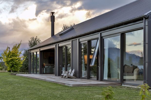 Exterior view of a contemporary single-storey home with a steep pitched metal roof, dark vertical metal cladding, extensive floor-to-ceiling glazing, and a timber deck opening onto a lawn. A chimney, outdoor chairs, and glimpses of the living interior are visible, with a dramatic cloudy sky and distant landscape beyond.