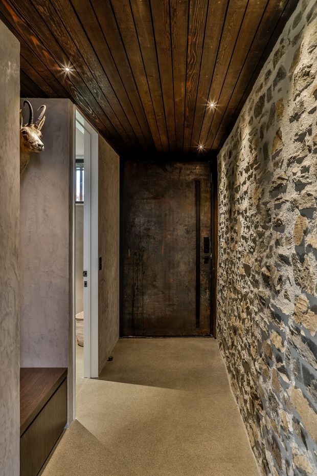 A narrow, atmospheric residential hallway with a rough exposed stone feature wall on the right, dark stained timber slat ceiling with recessed spotlights, warm concrete or polished plaster flooring, and a dark metal entrance door at the end. A partially visible built-in timber cabinet and textured plaster wall frame the left side, with an open doorway revealing another room. A decorative mounted animal head is visible on the wall.