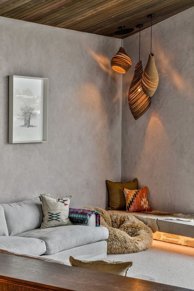 A cozy, textural living room with pale grey plastered walls, a warm timber slat ceiling, and sculptural woven pendant lights casting amber light. The room features a light grey sofa, a built-in window seat with earthy patterned cushions and a sheepskin throw, and a low timber coffee table. The muted natural palette and layered textiles create a relaxed, organic atmosphere.