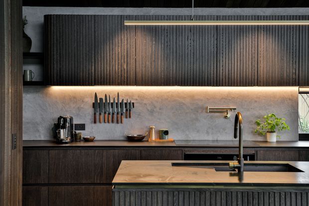 A sophisticated contemporary kitchen viewed straight on, featuring dark vertically fluted cabinetry, a warm grey textured stone or plaster splashback, integrated under-cabinet lighting, a timber-toned island benchtop, and a black undermount sink with a tall brushed brass tap. A knife rack, small appliances, open shelving, and a potted plant add functional detail. The lighting is warm and atmospheric, with strong linear illumination emphasizing the cabinetry texture.