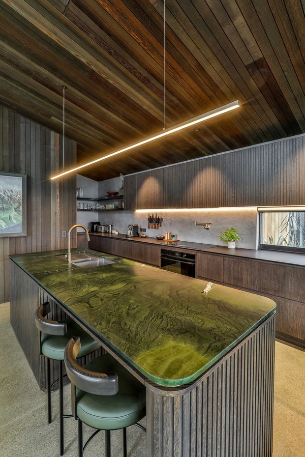 A contemporary kitchen dominated by dark timber finishes, with a large green-veined stone island and integrated seating. The room features vertical fluted timber cabinetry, a matching timber-lined ceiling, concrete-look walls, stainless-steel sink and appliances, open shelving, and linear pendant and under-cabinet lighting.