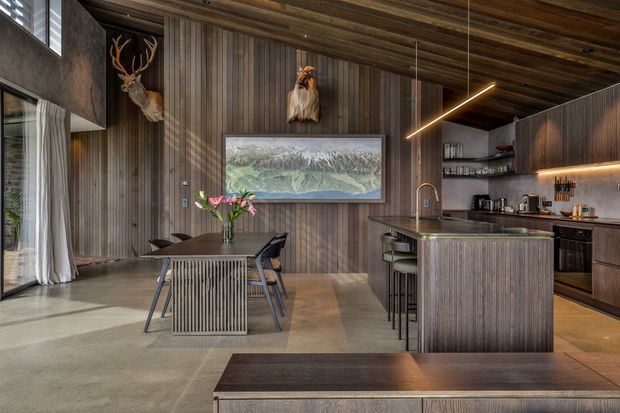 Open-plan dining and kitchen interior with dark vertical timber wall panelling, a matching timber-lined ceiling, concrete flooring, and a large island with integrated seating. The space features dark wood cabinetry, warm under-cabinet lighting, a linear pendant light, a dining table, and rustic antler wall decor. A high window and expansive glazing create a connection to the mountainous landscape outside.