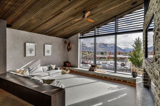 A contemporary living room with expansive floor-to-ceiling glazing and horizontal louvres overlooking a town and mountainous landscape. The room features a grey sectional sofa, built-in window seat, dark timber cabinetry, a textured concrete-look wall, exposed stonework, and a warm timber-lined ceiling with recessed lighting and a ceiling fan.