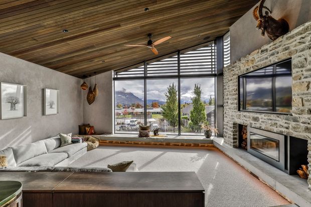 A warm, rustic-modern living room with a large corner window overlooking a mountainous suburban landscape. The room features a full-height rough stone fireplace wall with built-in fireboxes and television, a light sectional sofa, dark timber cabinetry, a wood-slat ceiling, concrete-textured walls, and a ceiling fan. Natural light fills the spacious room.