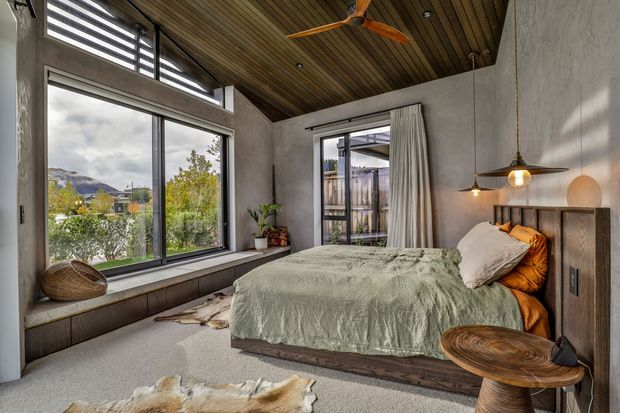 A warm, modern bedroom with a low platform bed, textured sage-green bedding, timber bedside table, and pendant lighting. The room features large black-framed windows overlooking a landscaped residential setting and hills, a wood-lined ceiling with a ceiling fan, pale textured walls, and a built-in window seat. Natural materials and muted earthy tones create a calm, inviting atmosphere.