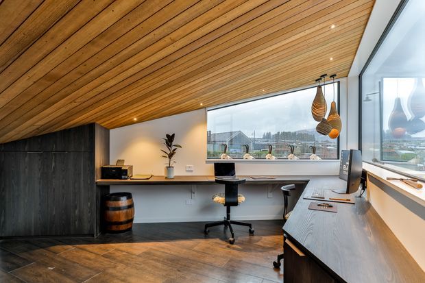 A contemporary home office with a built-in L-shaped timber desk, ergonomic stools, computer workstation, and integrated shelving. The room features a dramatic sloped timber-lined ceiling with recessed spotlights, dark vertical wall panelling and cabinetry, warm wood flooring, and large corner glazing overlooking an outdoor landscape. Woven pendant lights add a sculptural decorative element.