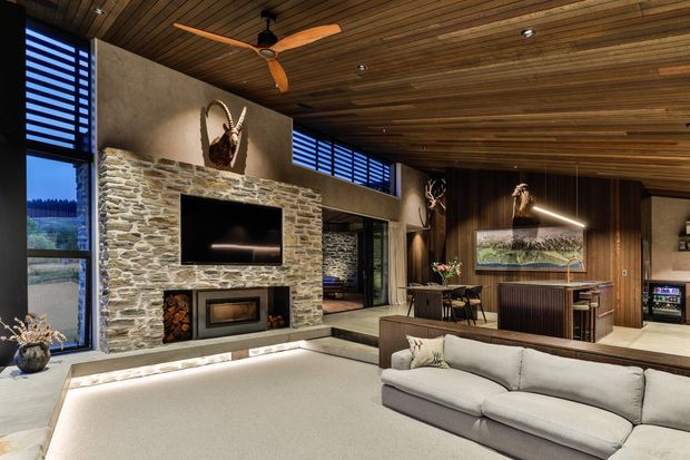 A spacious, warm-toned living room with a large stone fireplace wall, integrated television and fireplace, light sectional sofa, and timber-lined ceiling and walls. The room includes expansive dark-framed glazing with horizontal louvres, built-in timber cabinetry, a dining area, and a compact bar or kitchenette zone. Sculptural antler decor, timber flooring details, and recessed lighting contribute to a rustic yet refined lodge-like interior.