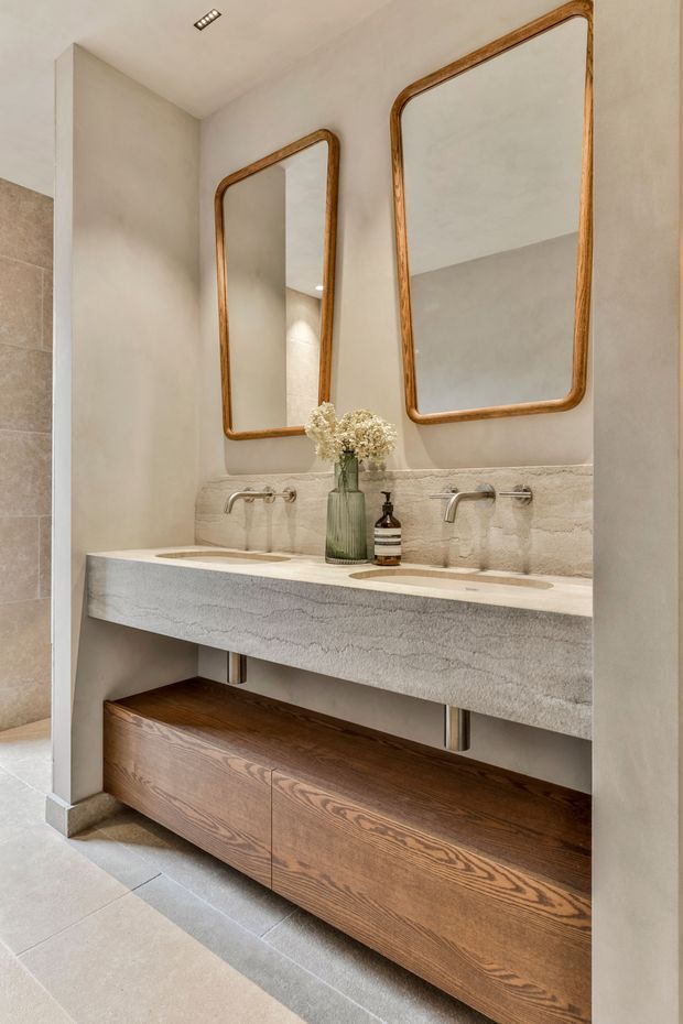 A refined contemporary bathroom featuring a long double vanity with an integrated pale stone basin and matching stone splashback. Two wall-mounted brushed-metal faucets sit beneath large rounded-rectangular mirrors with warm timber frames. The floating vanity includes a substantial natural timber drawer unit, while light stone or porcelain floor and wall surfaces create a warm neutral palette. A recessed ceiling spotlight and understated fixtures complete the space.